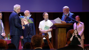 Kadr z transmisji wręczenia nagrody naukowej prof. Czaplińskiemu - na scenie przy mównicy przewodniczący rady miasta odczytuje werdykt jury, obok niego stoi prof. Czapliński trzymając swoją książkę, dalej prezydent Poznania z bukietem kwiatów i urzędnicy miejscy.