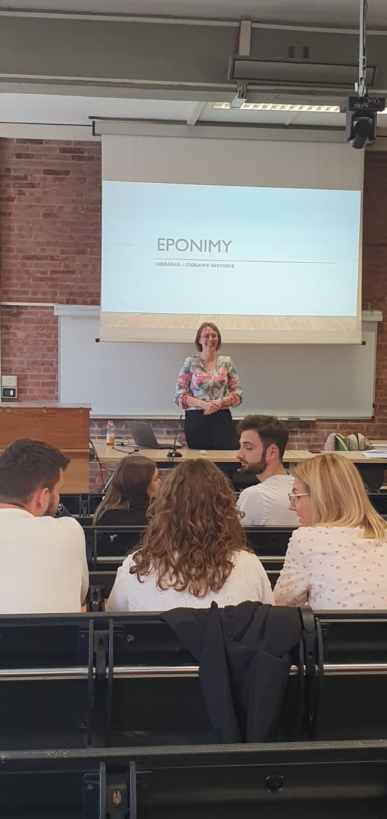 Młoda kobieta stoi w sali wykładowej, przed nią siedzą w rzędach studenci, za nią wyświetla się prezentacja z dużym napisem 