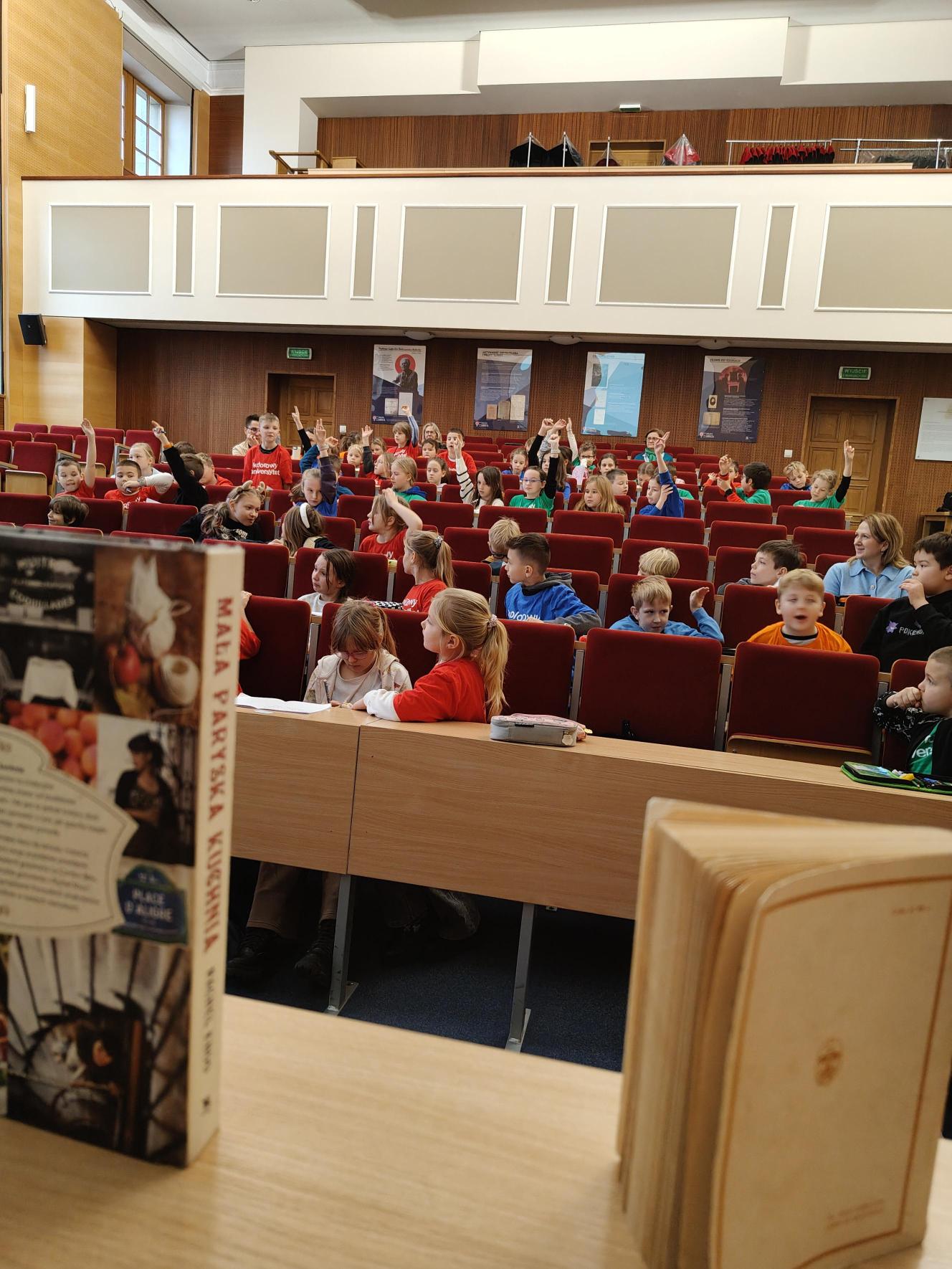 Dzieci w kolorowych koszulkach siedzą na fotelach ustawionych rzędami od góry do dołu. Na pierwszym planie widać biurko ze stojącymi na nim pionowo książkami.