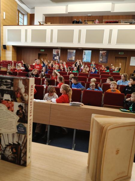 Dzieci w kolorowych koszulkach siedzą na fotelach ustawionych rzędami od góry do dołu. Na pierwszym planie widać biurko ze stojącymi na nim pionowo książkami.