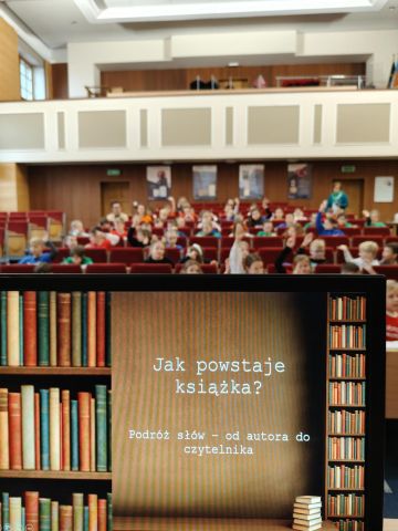Dzieci w kolorowych koszulkach siedzą na fotelach ustawionych rzędami od góry do dołu. Na pierwszym planie widać ekran z tytułem warsztatów.