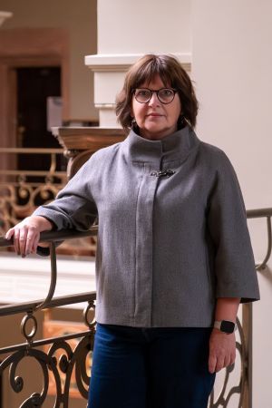 Kobieta w średnim wieku, w szarym golfie, ciemnych spodniach, z dużym wisiorkiem i w okularach stoi na drugim piętrze Collegium Maius lekko oparta ręką o zdobioną, złotawą balustradę.