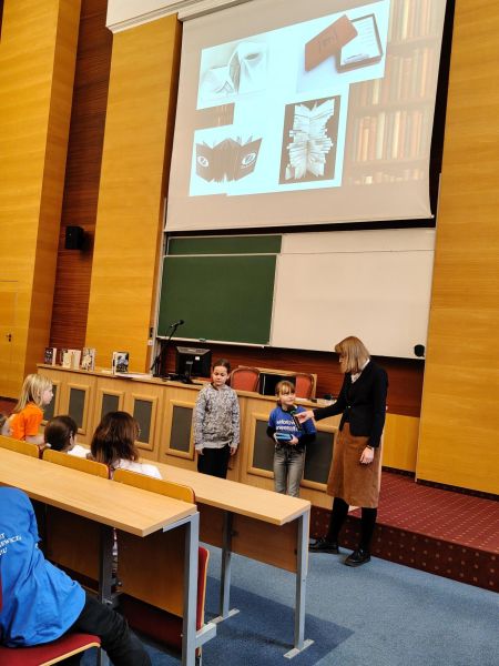 Dwoje dzieci stoi przy stole prezydialnym, jedno z nich odpowiada na zadane pytanie do mikrofonu trzymanego przez prowadzącą.