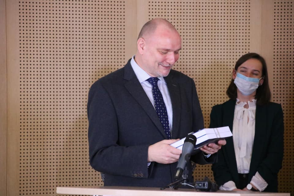 Zdjęcie z jubileuszu w Salonie Mickiewicza. Prof. Tomasz Lisowski przemawia przy mównicy, w dłoniach trzyma książkę, patrzy na tył jej okładki. Z tyłu widać dr Paulinę Michalską-Górecką