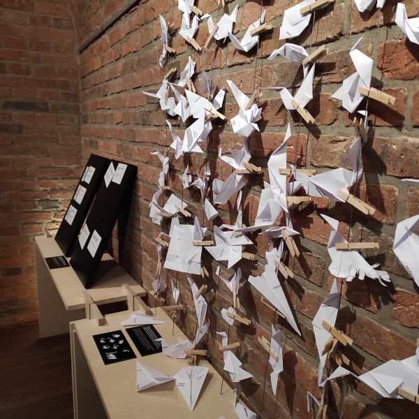 Na tle ceglanej ściany widać przywieszone papierowe samoloty i ptaki z origami.