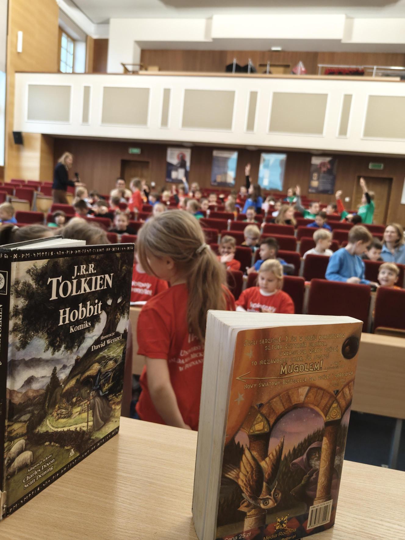 Dzieci w kolorowych koszulkach siedzą na fotelach ustawionych rzędami od góry do dołu. Na pierwszym planie widać biurko ze stojącymi na nim pionowo książkami.