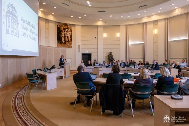 Zdjęcie przedstawia dużą salę konferencyjną z wysokim sufitem i jasnymi ścianami. Uczestnicy siedzą przy stołach ustawionych w podkowę, zwróceni w stronę mównicy i ekranu projekcyjnego, na którym wyświetlone jest logo Wydziału Filologii Polskiej i Klasycznej UAM. Przy mównicy stoi osoba prowadząca wystąpienie. W tle widoczna jest rzeźba postaci oraz duży obraz ścienny.