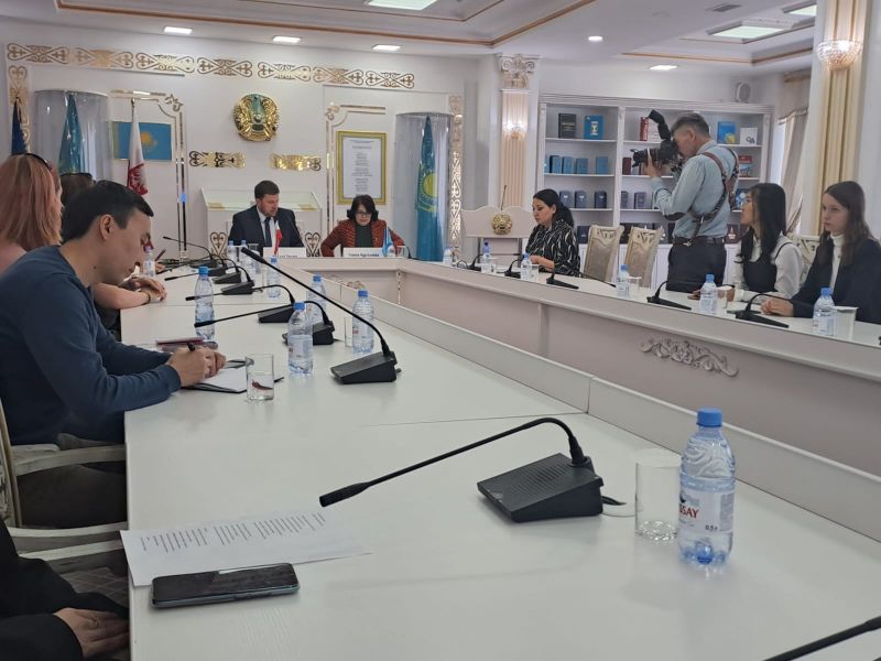 Biała, bogato zdobiona sala konferencyjna z długim białym stołem, na którym stoją mikrofony i butelki z wodą. Przy stole siedzi kilkanaście osób, w tle widać flagi Kazachstanu.