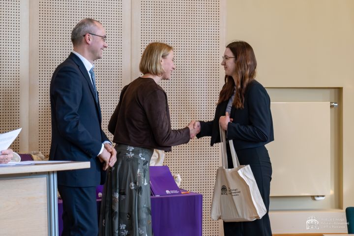 Uroczyste wręczenie nagrody w sali konferencyjnej. Osoba stojąca po prawej stronie odbiera upominek w płóciennej torbie i podaje rękę osobie znajdującej się pośrodku. Obok stoi kolejna osoba w eleganckim stroju. W tle widoczne są fioletowe pudełka ustawione na stole oraz perforowana ściana panelowa.