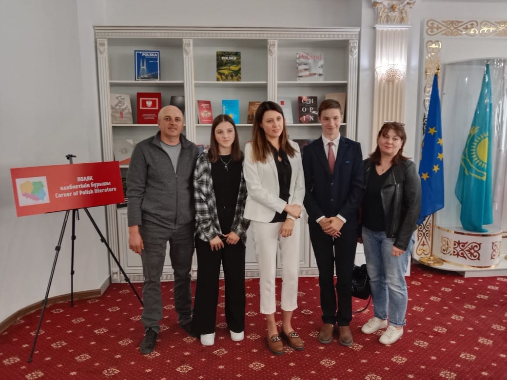 5 osób stoi w białym pomieszczeniu wyłożonym czerwonym dywanem w złote wzory, pozują do zdjęcia robionego przez osobę obok, której nie widać. Obok nich stoją flagi Unii Europejskiej i Kazachstanu. Za nimi przeszklona witryna z książkami.
