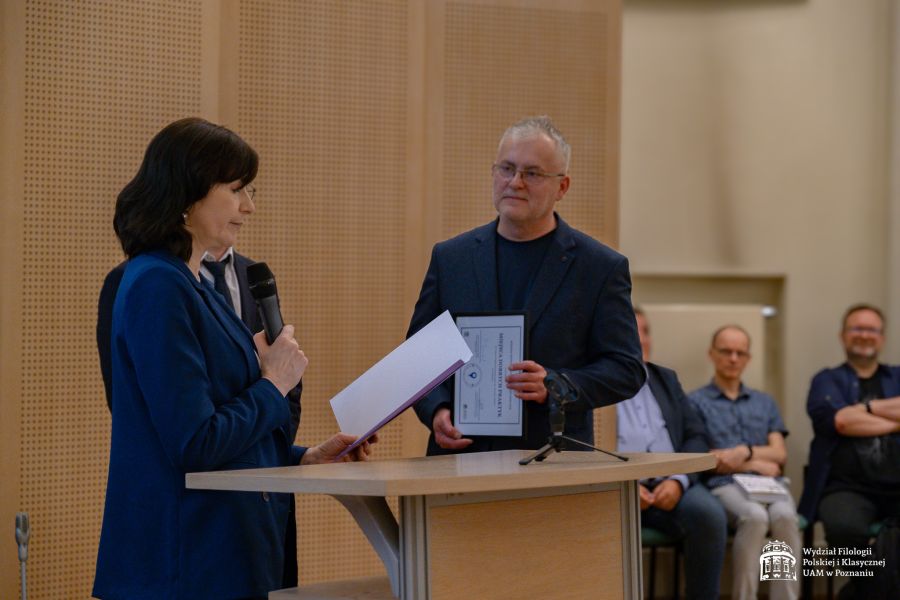 Prof. Elżbieta Winiecka odczytuje przy mównicy laudację, dalej stoi mężczyzna w średnim wieku, trzymający w dłoniach oprawiony w ramkę dyplom.