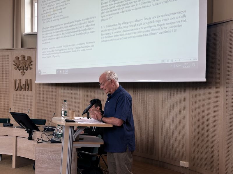 W Salonie Mickiewicza przy mównicy stoi starszy mężczyzna w koszuli, przemawia do mikrofonu, na rzutniku wyświetla się prezentacja z dużą ilością tekstu.