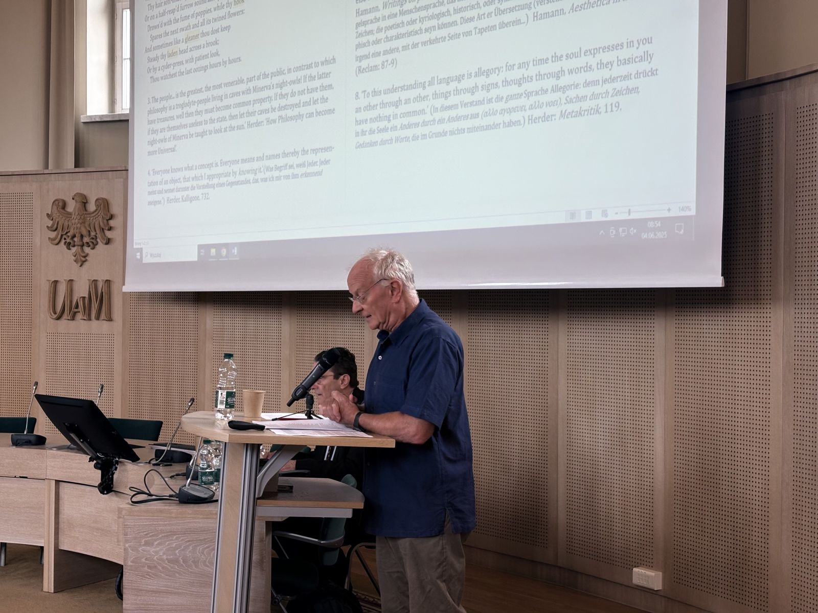 W Salonie Mickiewicza przy mównicy stoi starszy mężczyzna w koszuli, przemawia do mikrofonu, na rzutniku wyświetla się prezentacja z dużą ilością tekstu.