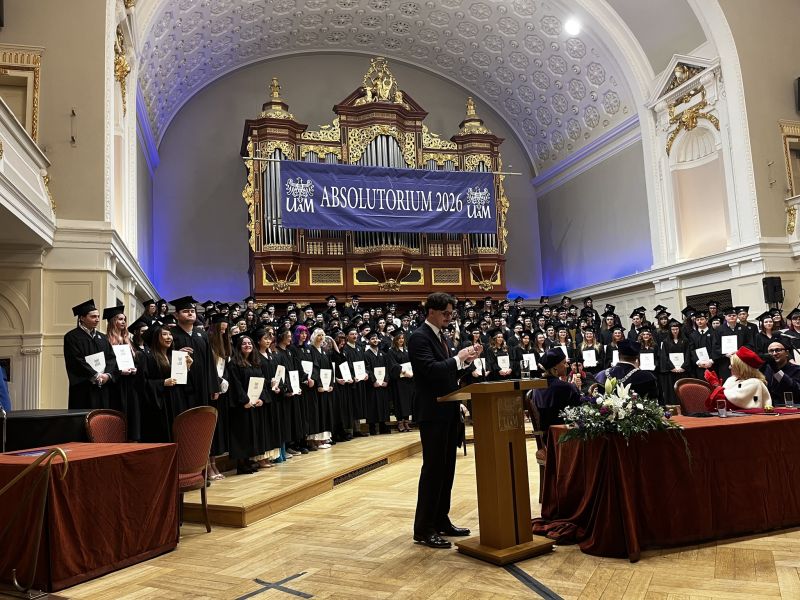 Uroczystość absolutorium UAM 2026 w ozdobnej auli z organami i bogatym wystrojem. Na scenie stoi grupa absolwentów w togach z dyplomami, nad którym widoczny jest baner wydarzenia. Przy mównicy trwa przemówienie.