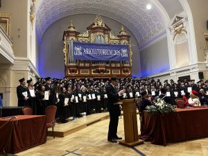 Uroczystość absolutorium UAM 2026 w ozdobnej auli z organami i bogatym wystrojem. Na scenie stoi grupa absolwentów w togach z dyplomami, nad którym widoczny jest baner wydarzenia. Przy mównicy trwa przemówienie.