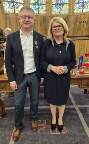 Fotografia przedstawia kobietę i mężczyznę w średnim wieku, którzy pozują w eleganckim wnętrzu. Mężczyzna ma przypięty medal do marynarki. W tle drewniane meble i witrażowe okna. 