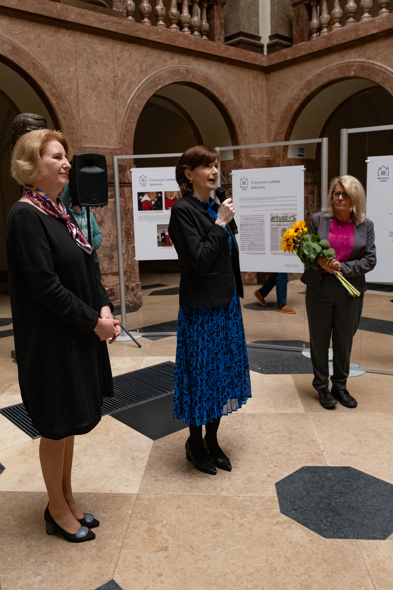 W holu głównym Collegium Maius, pośród tablic z wystawą o Wisławie Szymborskiej, stoją od lewej: prof. dr hab. Joanna Wójcik, prof. dr hab. Elżbieta Winiecka i prof. dr hab. Bogumiła Kaniewska, Rektor UAM. Pani Rektor trzyma w ręku bukiet słoneczników.