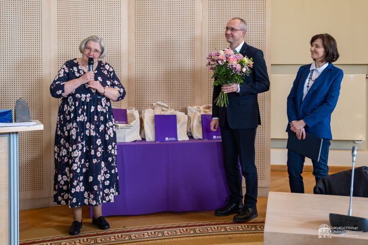 W jasnej sali trzy osoby stoją przed stołem nakrytym fioletowym obrusem, na którym ustawiono torby i pudełka upominkowe. Kobieta po lewej stronie trzyma mikrofon i stoi w kwiecistej sukience. Mężczyzna w garniturze stojący pośrodku trzyma bukiet kwiatów. Po prawej stronie stoi kobieta w garniturze, trzymająca teczkę. W tle widoczna jest perforowana ściana oraz elementy wyposażenia sali konferencyjnej.