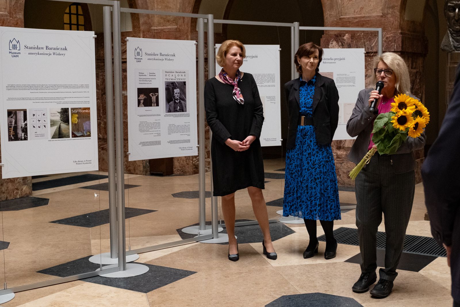 W holu głównym Collegium Maius na tle tablic z wystawą o Wisławie Szymborskiej stoją: prof. dr hab. Joanna Wójcik, prof. dr hab. Elżbieta Winiecka i Jej Magnificencja Rektor UAM prof. dr hab. Bogumiła Kaniewska. Pani Rektor przemawia do mikrofonu, w ręku trzyma bukiet słoneczników.