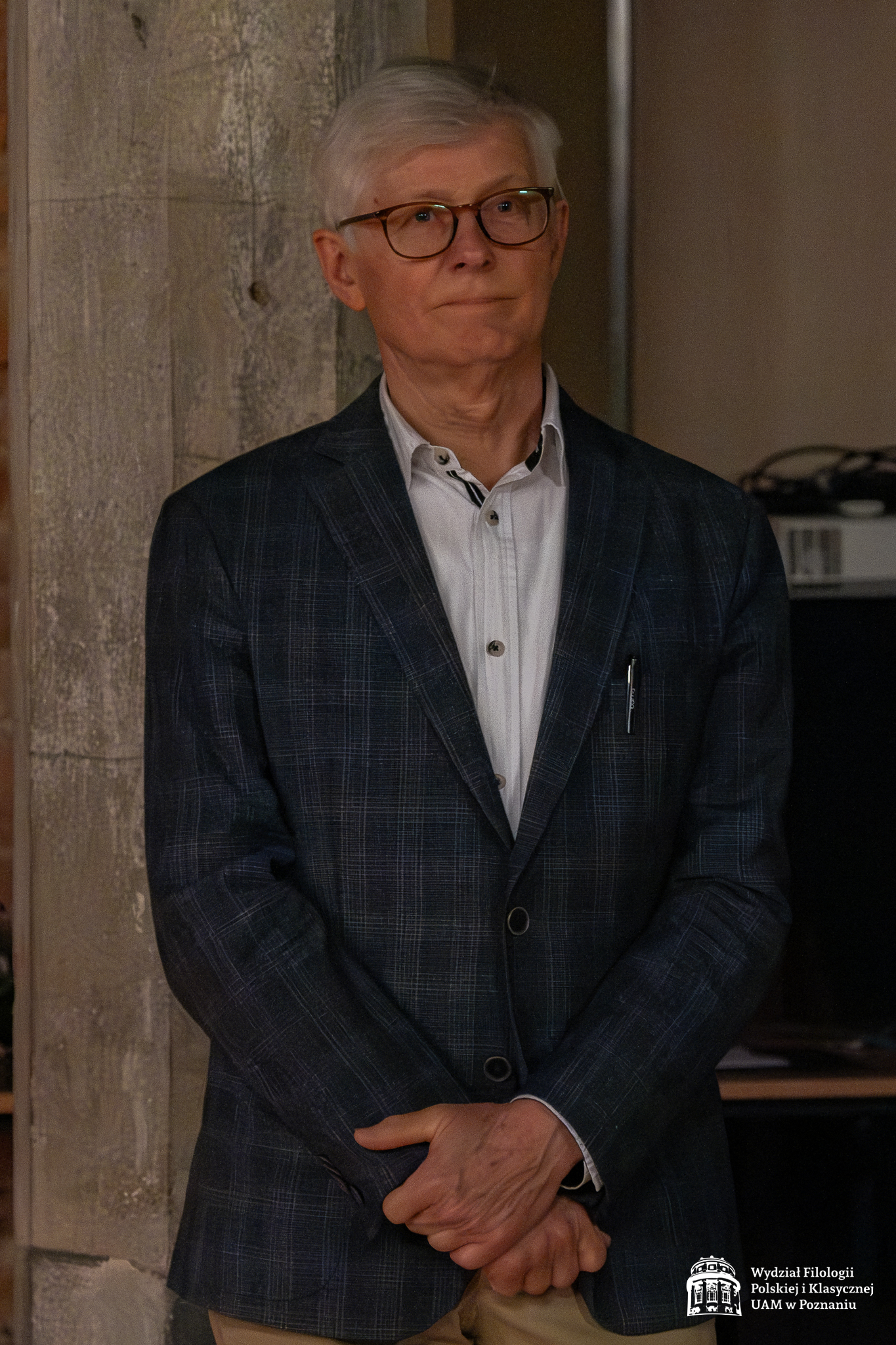 Prof. Zbigniew Przychodniak stoi w sali teatralnej, z poważną miną słuchając przemówienia.