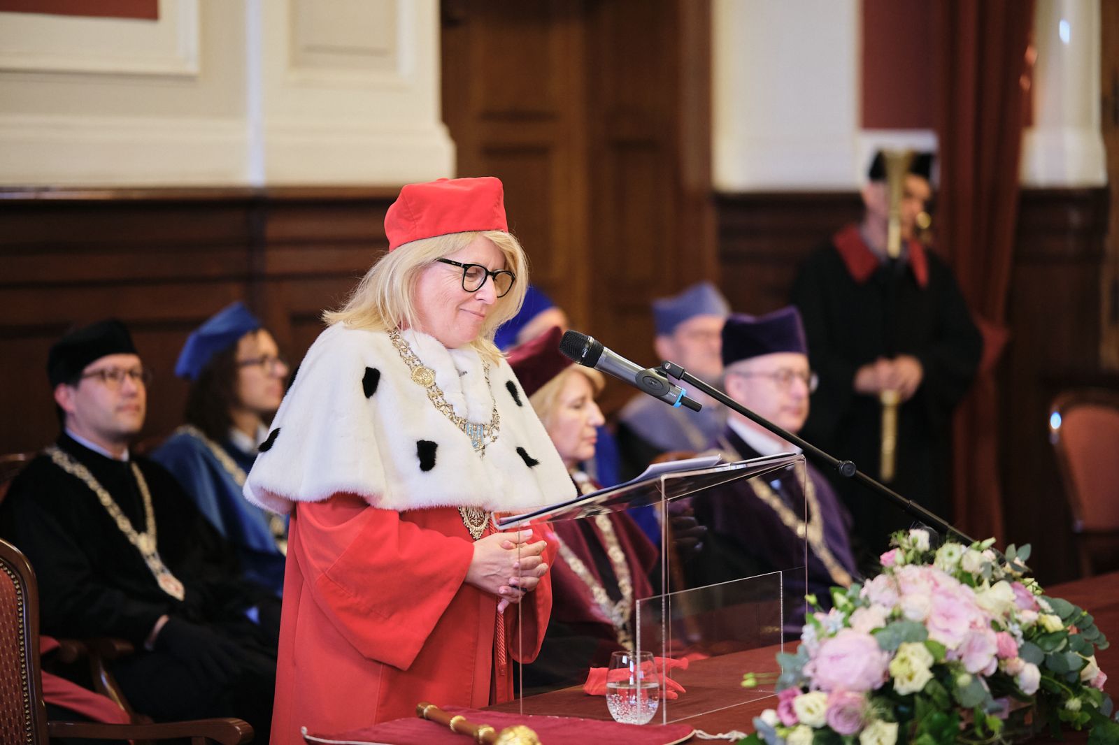 Jej Magnificencja Rektor Bogumiła Kaniewska, ubrana w płaszcz i biret rektorski, przemawia do mikrofonu w Auli Lubrańskiego.