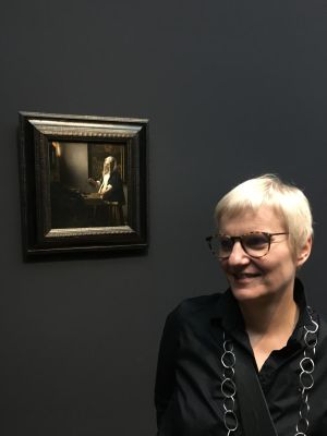 Prof. Agata Stankowska w ciemnej koszuli, okularach i krótkich blond włosach stoi obok obrazu Vermeera 