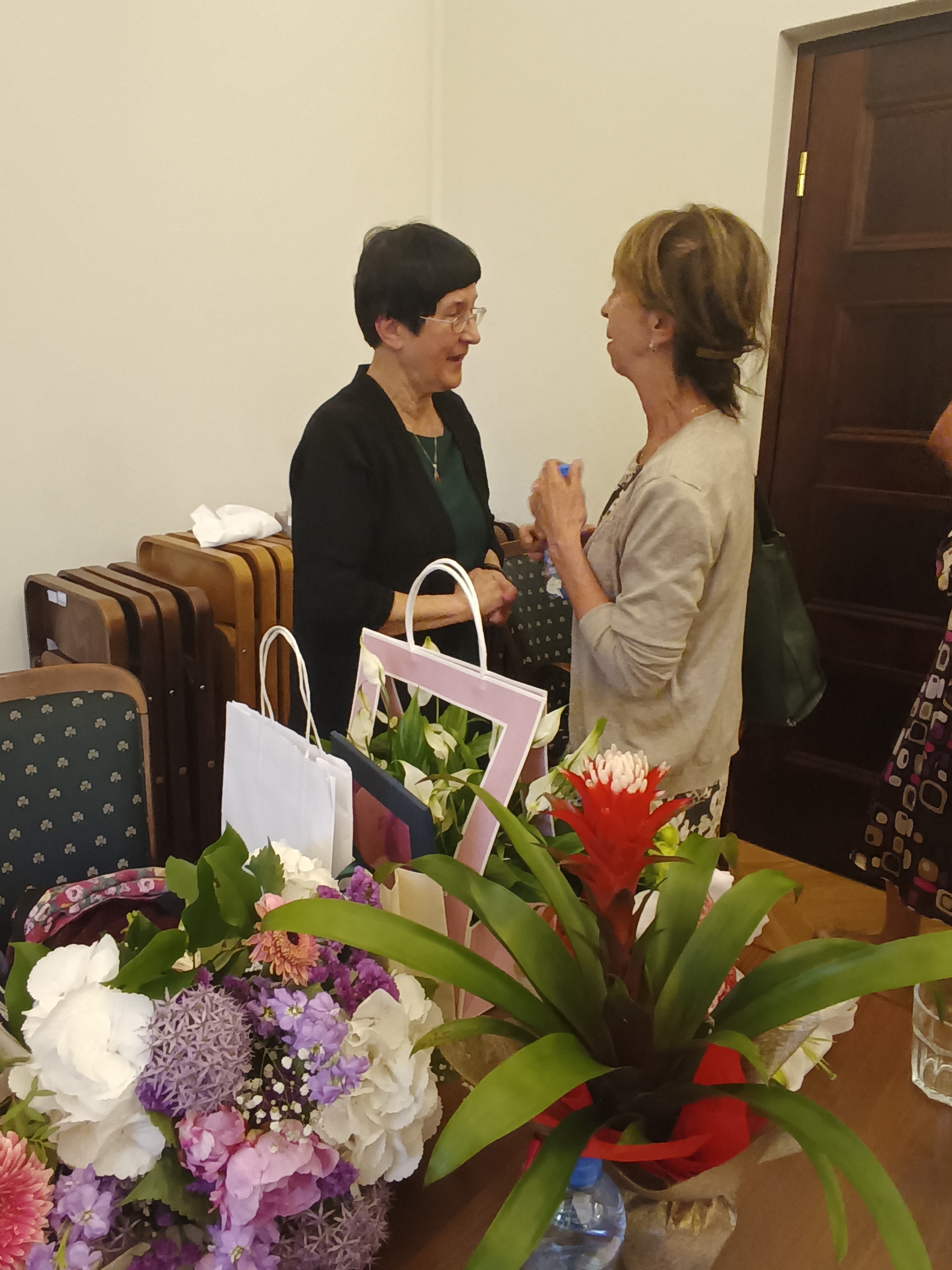 Prof. Elżbieta Nowicka składa gratulacje jubilatce, przed nimi na stole leży mnóstwo kwiatów i prezentów.