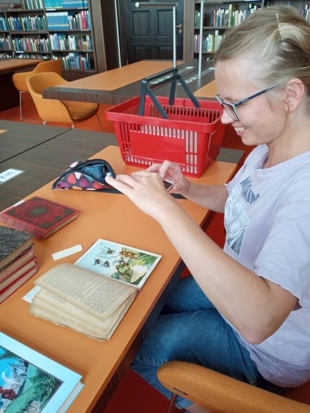 Młoda kobieta siedzi przy stole, przed nią leży otwarta książka z kolorową ilustracją, którą kobieta fotografuje, szeroko się uśmiechając. Wokół niej widać regały z kolejnymi książkami.