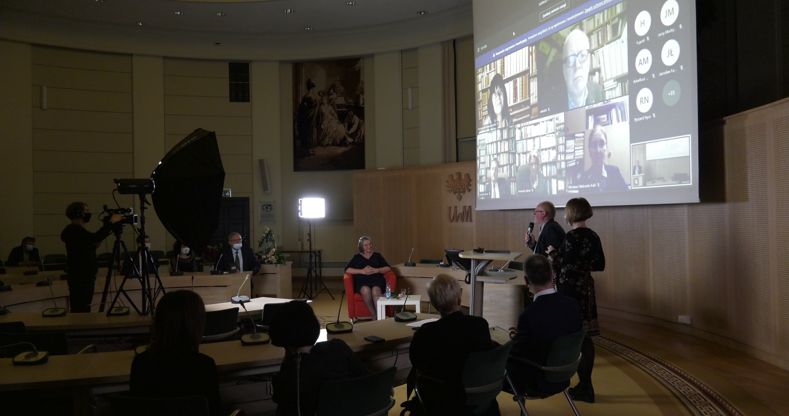 Rzut ogólny na Salon Mickiewicza - widać ekran, na którym widać twarze gości uczestniczących zdalnie, przy pulpicie stoi prof. Wojciech Otto i przemawia, widać również kamerzystę, oświetlenie i część publiki.