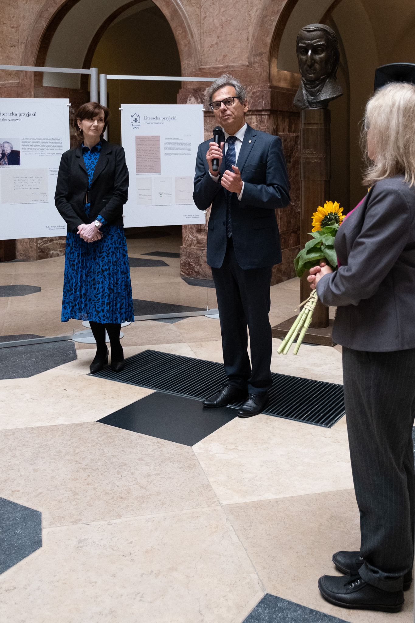 W holu głównym Collegium Maius na tle wystawy o Wisławie Szymborskiej stoją: prof. Elżbieta Winiecka, prof. Andrzej Gulczyński, dyrektor Muzeum UAM oraz JMR prof. Bogumiła Kaniewska. Prof. Gulczyński przemawia do mikrofonu.