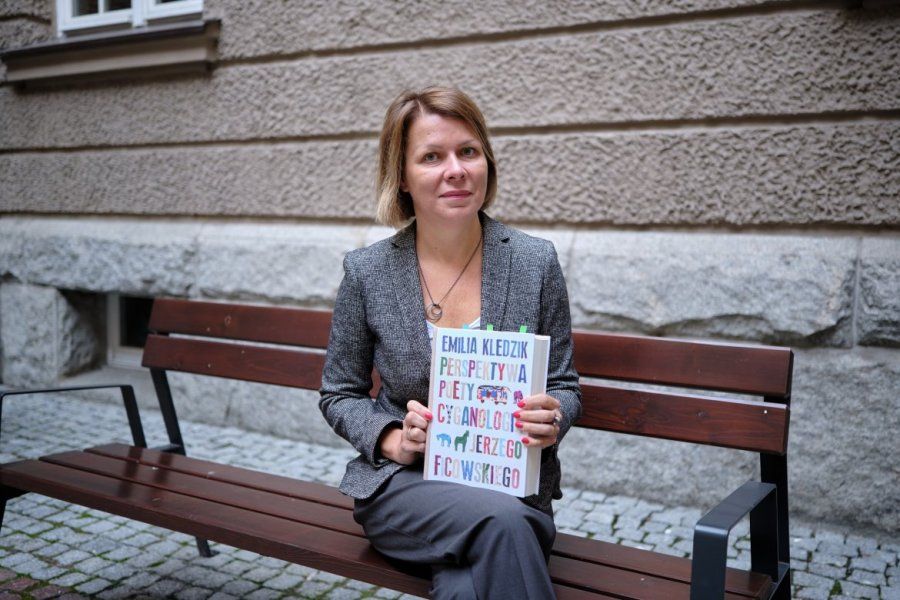 Dr hab. Emilia Kledzik siedzi na ławce na dziedzińcu Collegium Maius, w dłoniach trzyma swoją publikację 