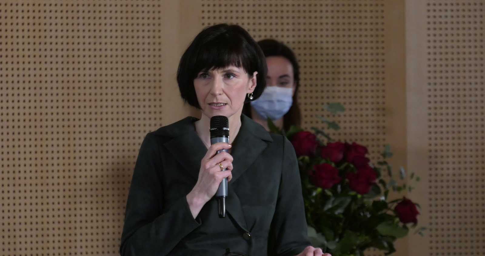 Prof. Elżbieta Winiecka przemawia do mikrofonu, za nią stoi dr Sylwia Karolak, w ręku trzyma bukiet czerwonych róż.
