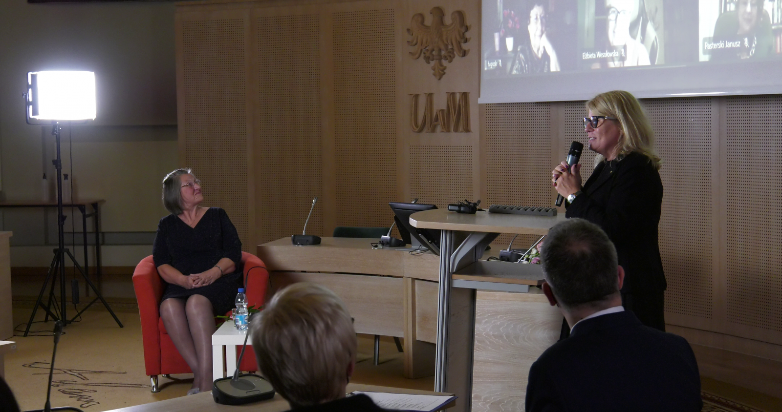 Rektor UAM prof. Bogumiła Kaniewska składa życzenia jubilatce, stojąc przy pulpicie w Salonie Mickiewicza. Na ekranie widać twarze gości uczestniczących w wydarzeniu zdalnie. Jubilatka siedzi w czerwonym fotelu, patrzy na ekran.
