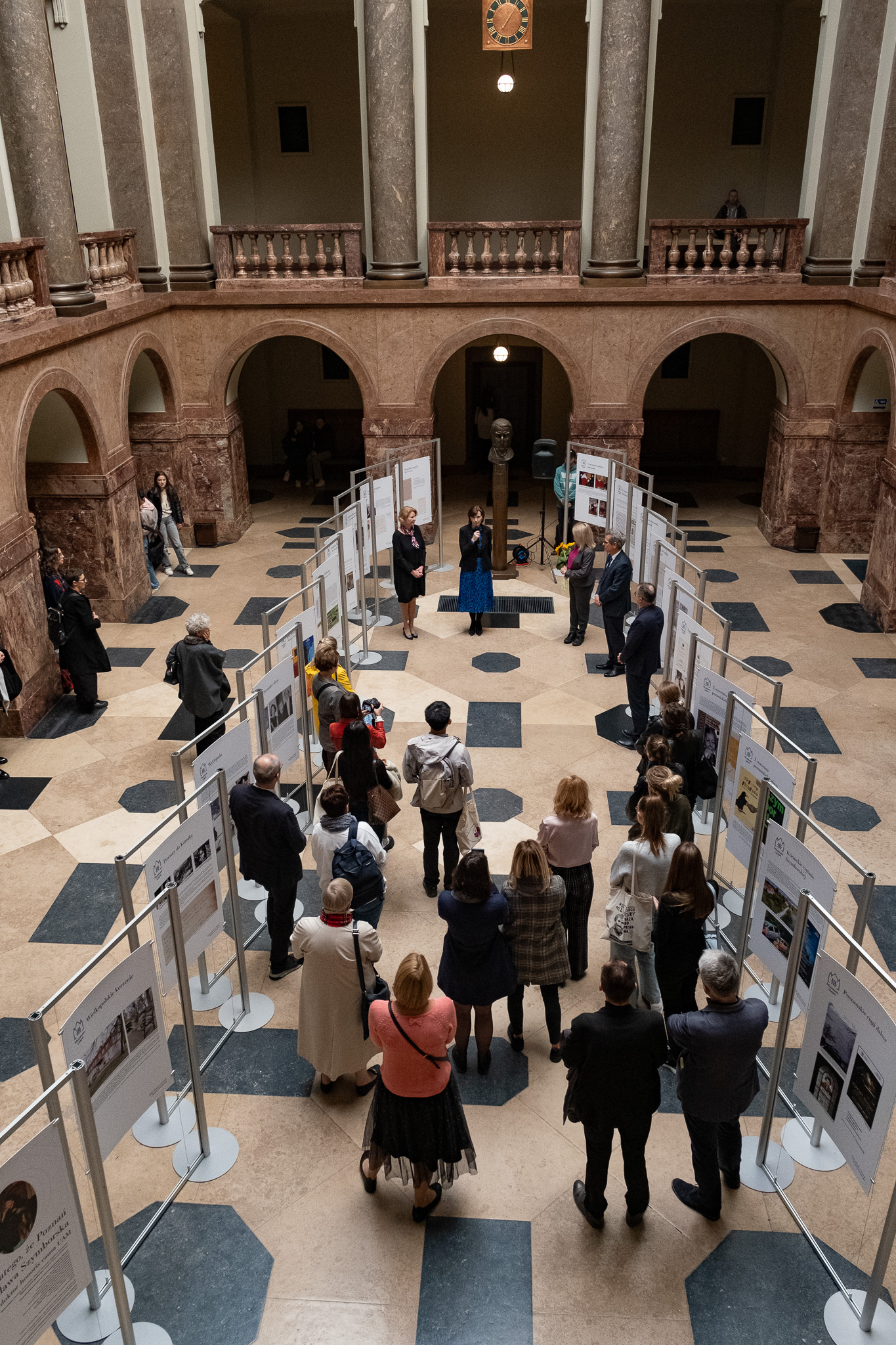 Zdjęcie holu głównego Collegium Maius z góry, widać plansze z wystawą o Wisławie Szymborskiej i stojących przed nimi uczestników otwarcia wystawy.