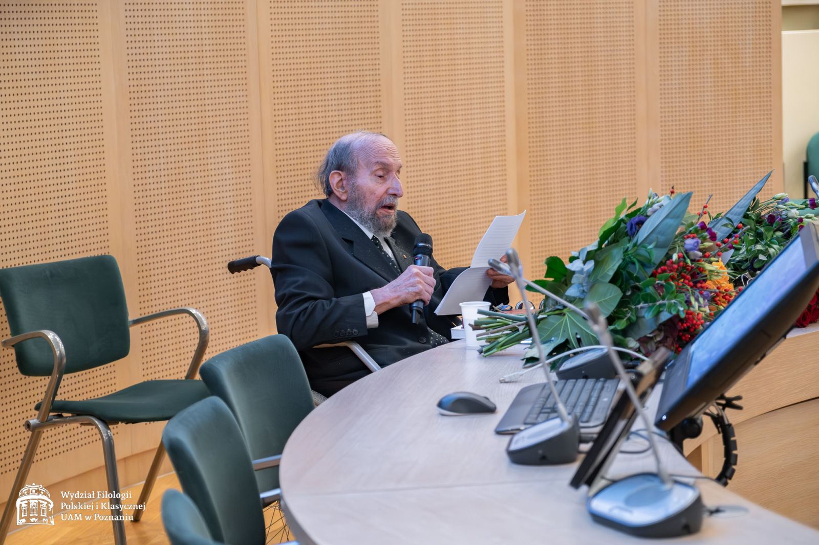 Profesor Tomasz Lewandowski siedzi w wózku inwalidzkim za stołem prezydialnym w Salonie Mickiewicza, przemawia do mikrofonu, posiłkując się kartką.