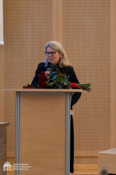 JM Rektor Bogumiła Kaniewska przemawia do mikrofonu, stojąc za mównicą, na której leży bukiet róż.