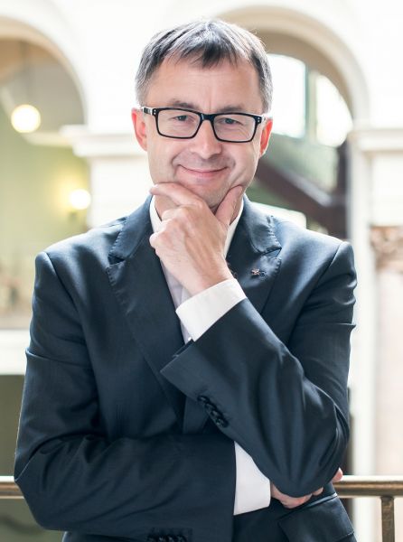 Zdjęcie portretowe prof. Tomasza Mizerkiewicza, na którym profesor ubrany w szary garnitur i krawat trzyma dłoń na brodzie, drugą ręką podpierając łokieć pierwszej, patrzy wprost w obiektyw przez okulary i uśmiecha się do widza.